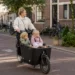 Ouder fietst met twee kinderen in een bakfiets door een rustige Nederlandse straat.